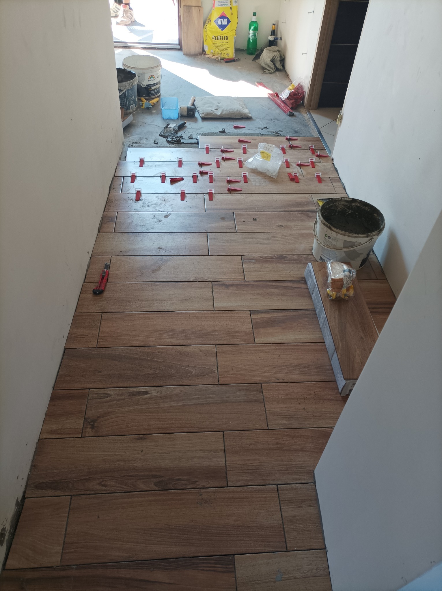 Układanie podłogi z płytek imitujących drewno w korytarzu, widoczne czerwone klipsy poziomujące, wiadro z zaprawą i nóż do tapet przy ścianie.