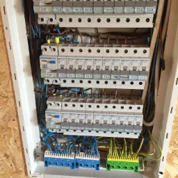 Rozdzielnica elektryczna z zainstalowanymi bezpiecznikami HAGER, widoczne przewody niebieskie, brązowe i żółto-zielone oraz listwy zaciskowe.