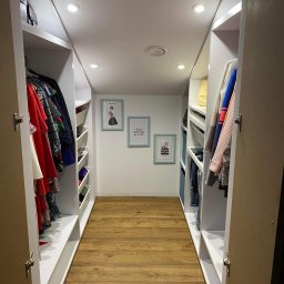 JS Perfect House - Jarosław Szymczak - Wnętrze garderoby z białymi szafami w zabudowie, drewnianą podłogą i dekoracyjnymi obrazkami na ścianie.