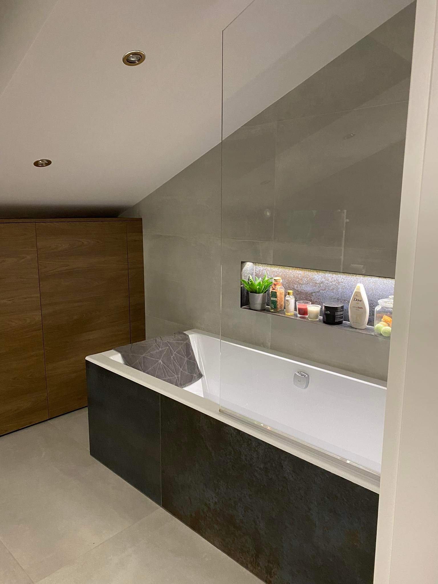 Nowoczesna łazienka na poddaszu z wanną obudowaną płytami imitującymi kamień, szklanym parawanem, wnęką z oświetleniem i drewnianą szafą w zabudowie.