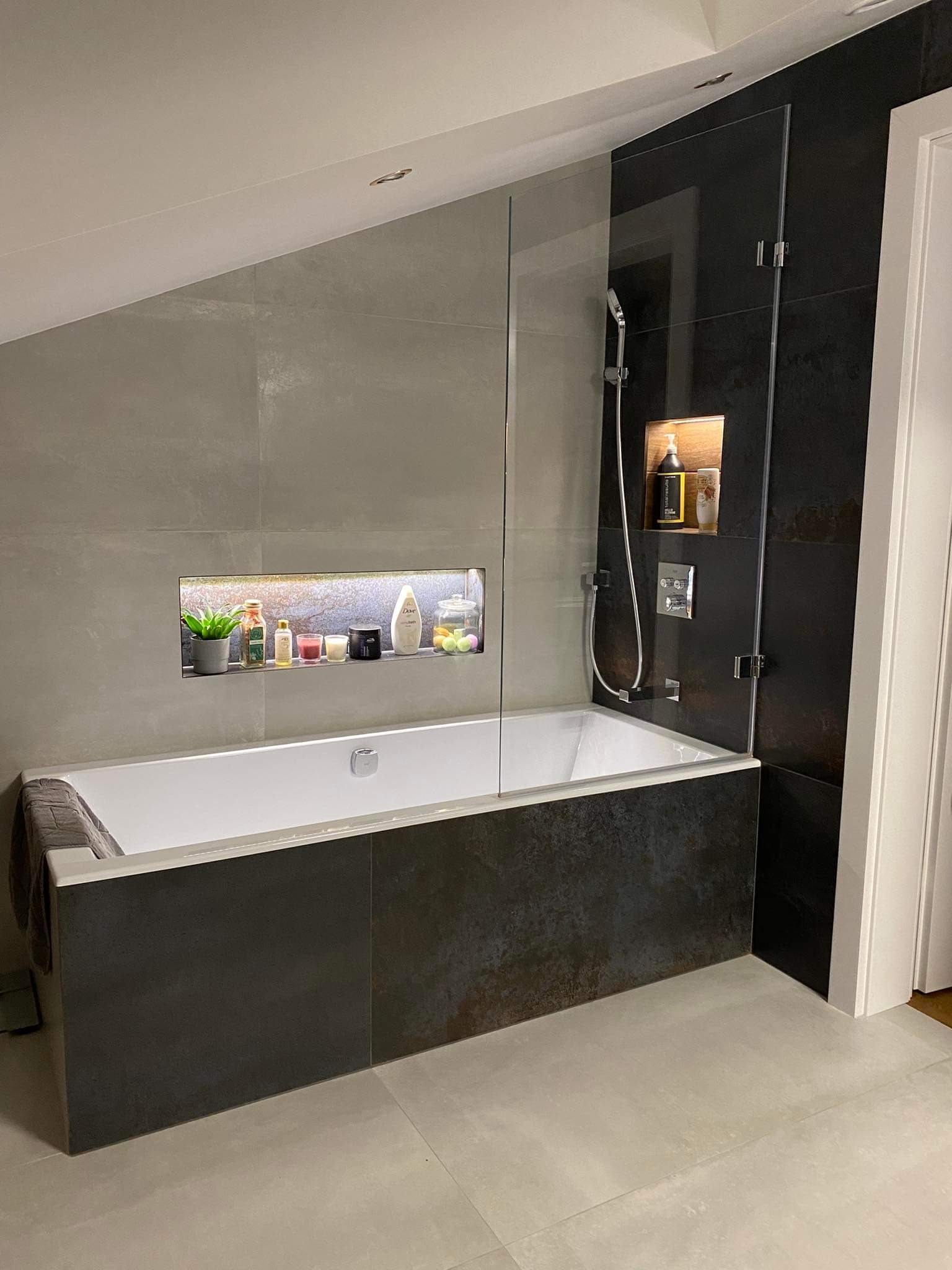 Nowoczesna łazienka z wanną obudowaną ciemnymi płytkami, przeszklona kabina prysznicowa i wnęki z oświetleniem eksponujące dekoracje i kosmetyki.