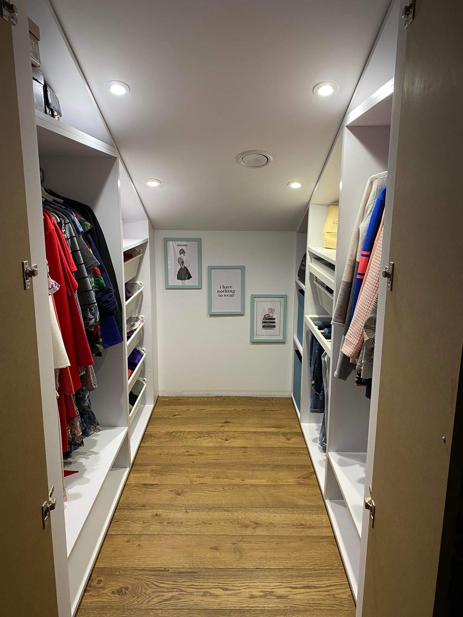 Wnętrze garderoby z białymi szafami w zabudowie, drewnianą podłogą i dekoracyjnymi obrazkami na ścianie.