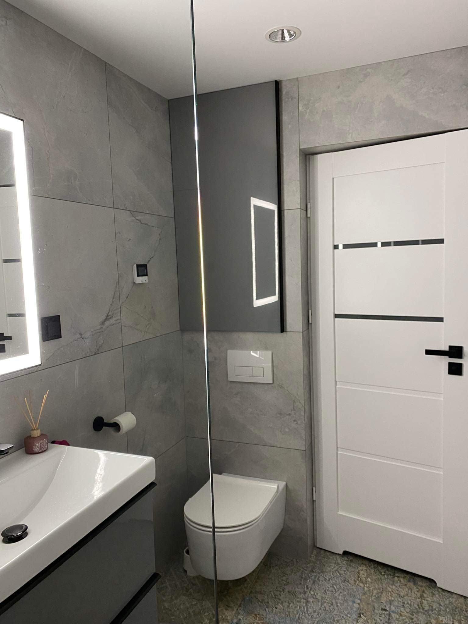 Nowoczesna łazienka z szarymi płytkami imitującymi marmur, białą umywalką z szarą szafką, podświetlanym lustrem, wiszącą toaletą i białą, minimalistyczną drzwiami z czarną klamką.