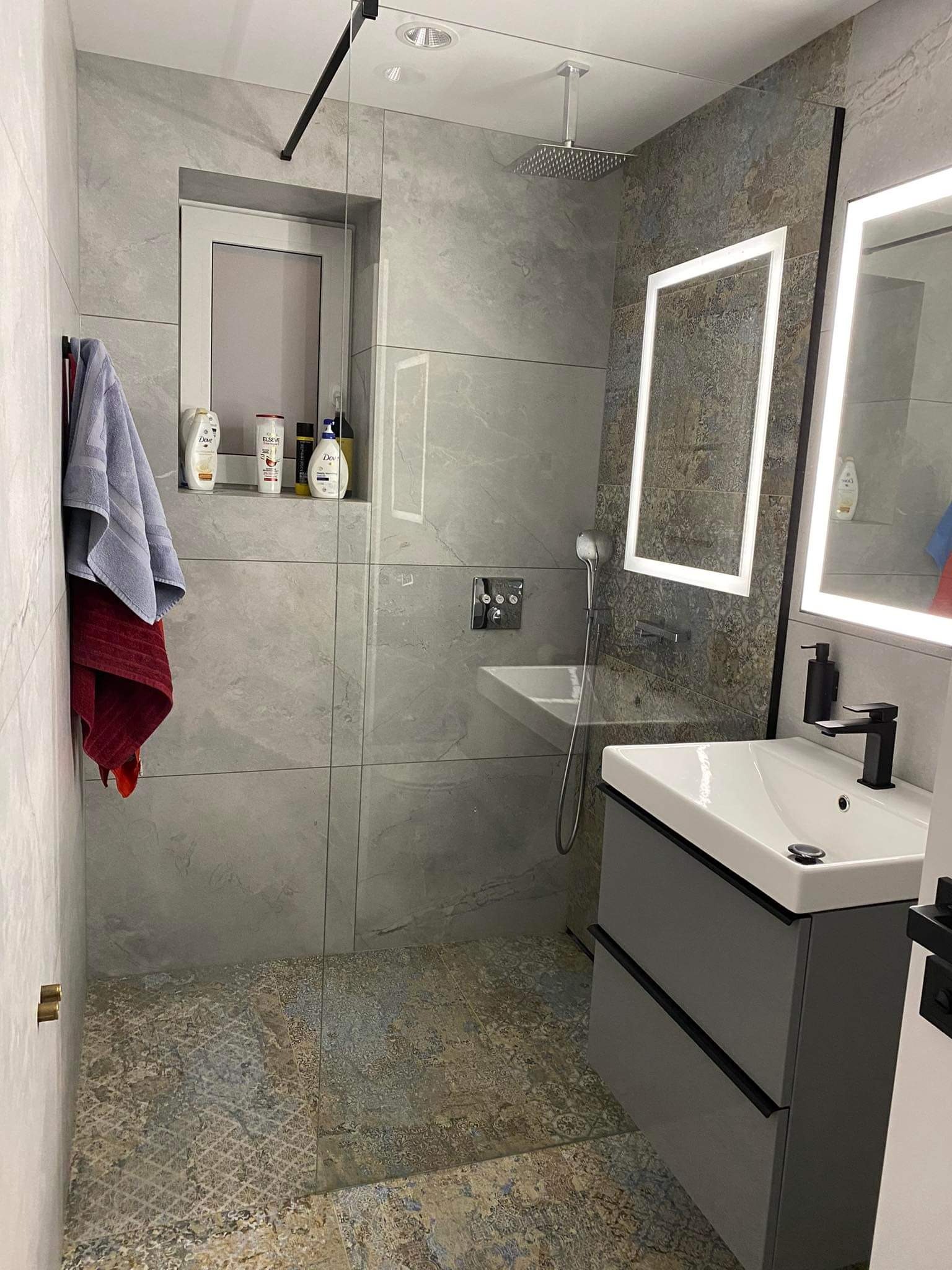 Nowoczesna łazienka z szarymi płytkami imitującymi kamień, kabiną prysznicową typu walk-in, umywalką z szafką w kolorze grafitowym i podświetlanymi lustrami.