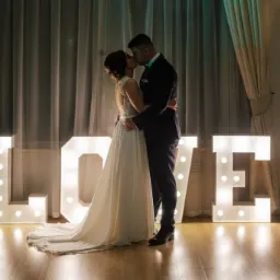 Pocałunek pary młodej na tle podświetlanych liter LOVE, suknia ślubna z koronkowymi plecami, ciemny garnitur pana młodego, kremowe zasłony w tle.