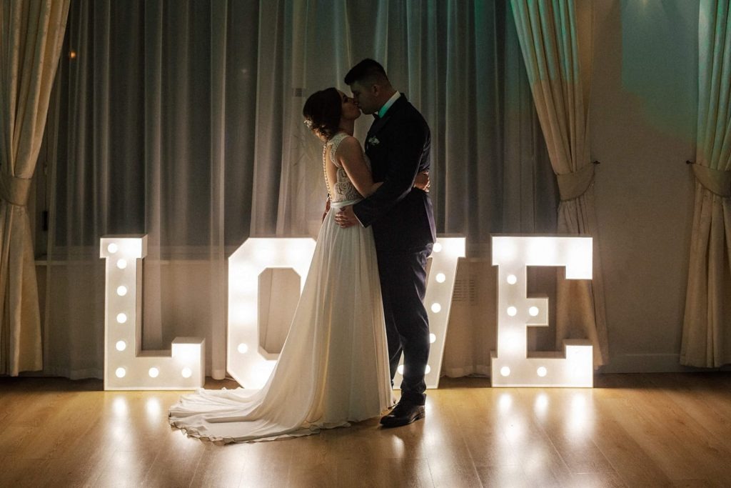 Pocałunek pary młodej na tle podświetlanych liter LOVE, suknia ślubna z koronkowymi plecami, ciemny garnitur pana młodego, kremowe zasłony w tle.