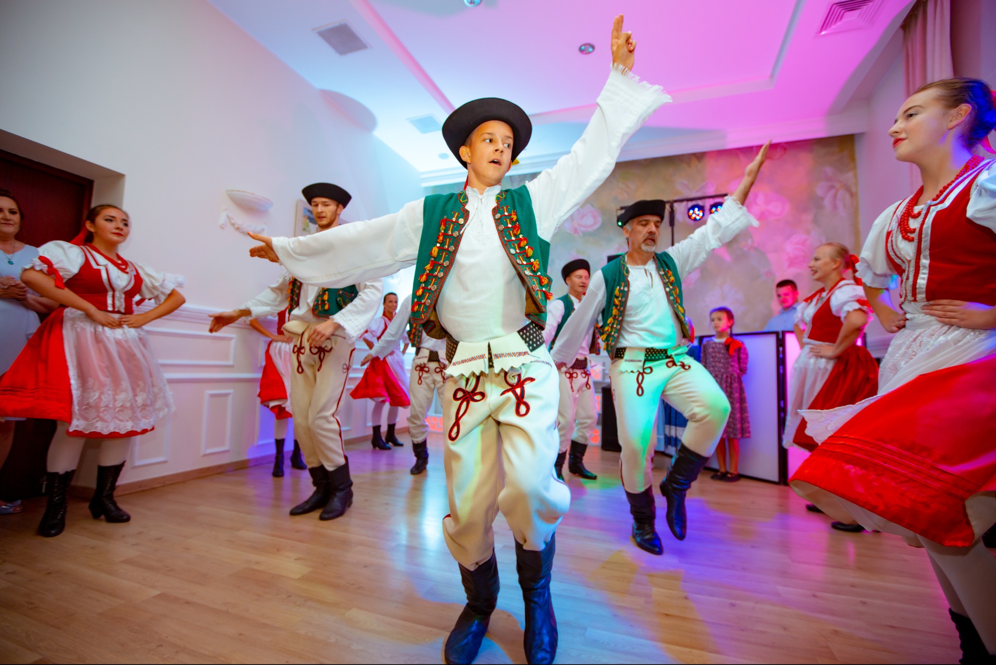 Zespół taneczny w tradycyjnych strojach ludowych prezentuje układ taneczny na scenie. W tle widoczny sprzęt nagłośnieniowy i oświetleniowy.