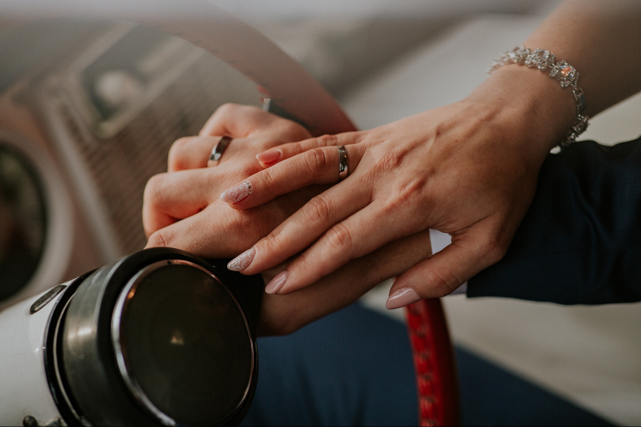 Dłonie nowożeńców z obrączkami na tle kierownicy klasycznego auta, bransoletka na nadgarstku panny młodej, delikatne zdobienia paznokci.