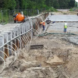 Widok na plac budowy z wykopem zabezpieczonym ściankami szczelnymi, systemem drenażowym, pracującymi robotnikami w kamizelkach odblaskowych i pomarańczową pompą odwadniającą oraz żółtą koparką.