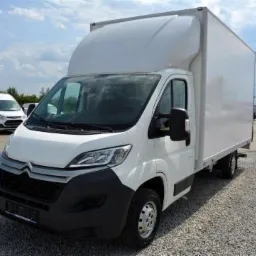 Biały samochód dostawczy Citroen Jumper z wysoką zabudową kontenerową na parkingu żwirowym, widoczne inne pojazdy w tle pod błękitnym niebem.