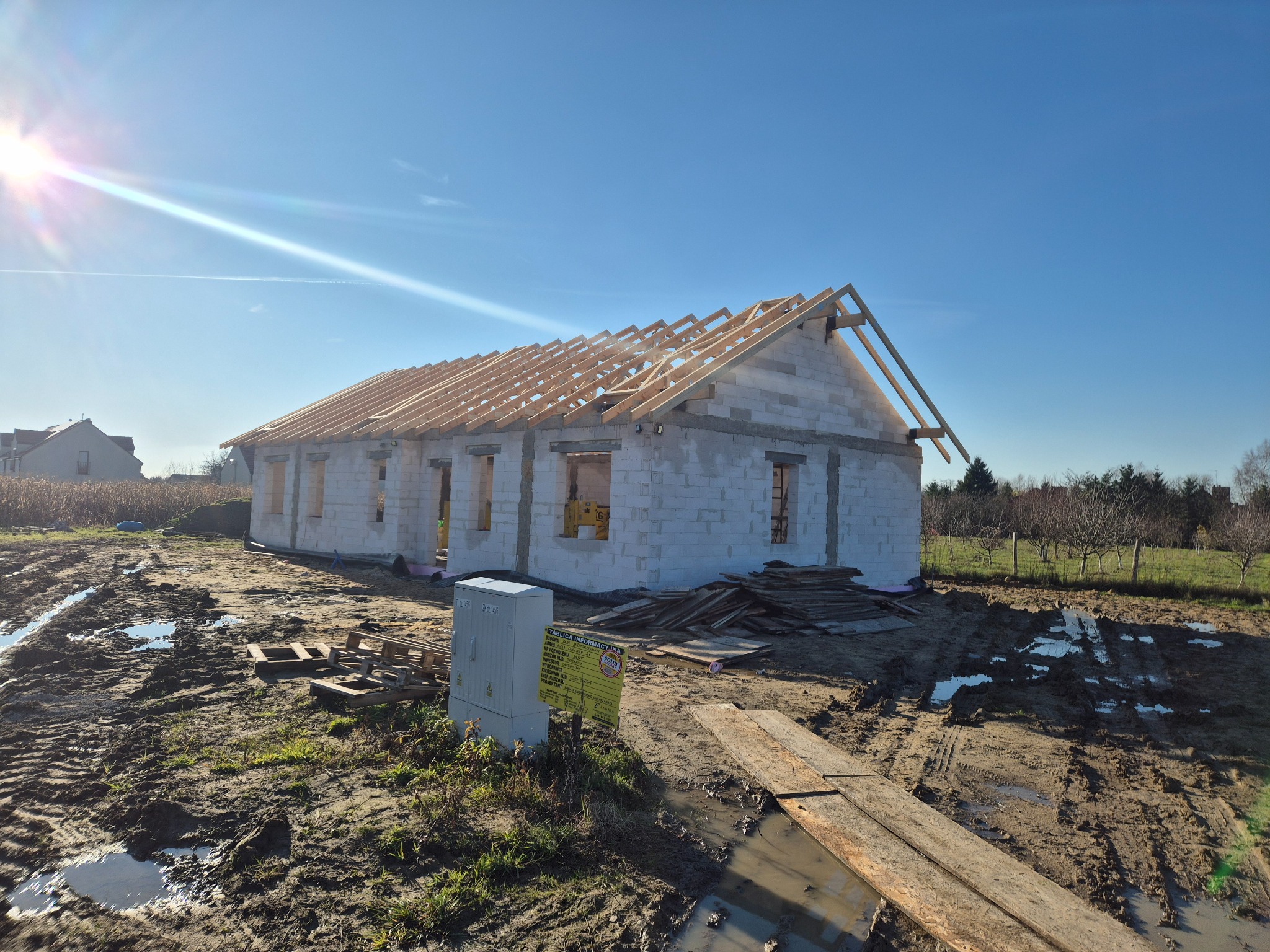 Budowa domu jednorodzinnego w stanie surowym otwartym, ściany z białego bloczka, widoczna więźba dachowa, tablica informacyjna budowy, teren budowy po deszczu, słoneczny dzień.