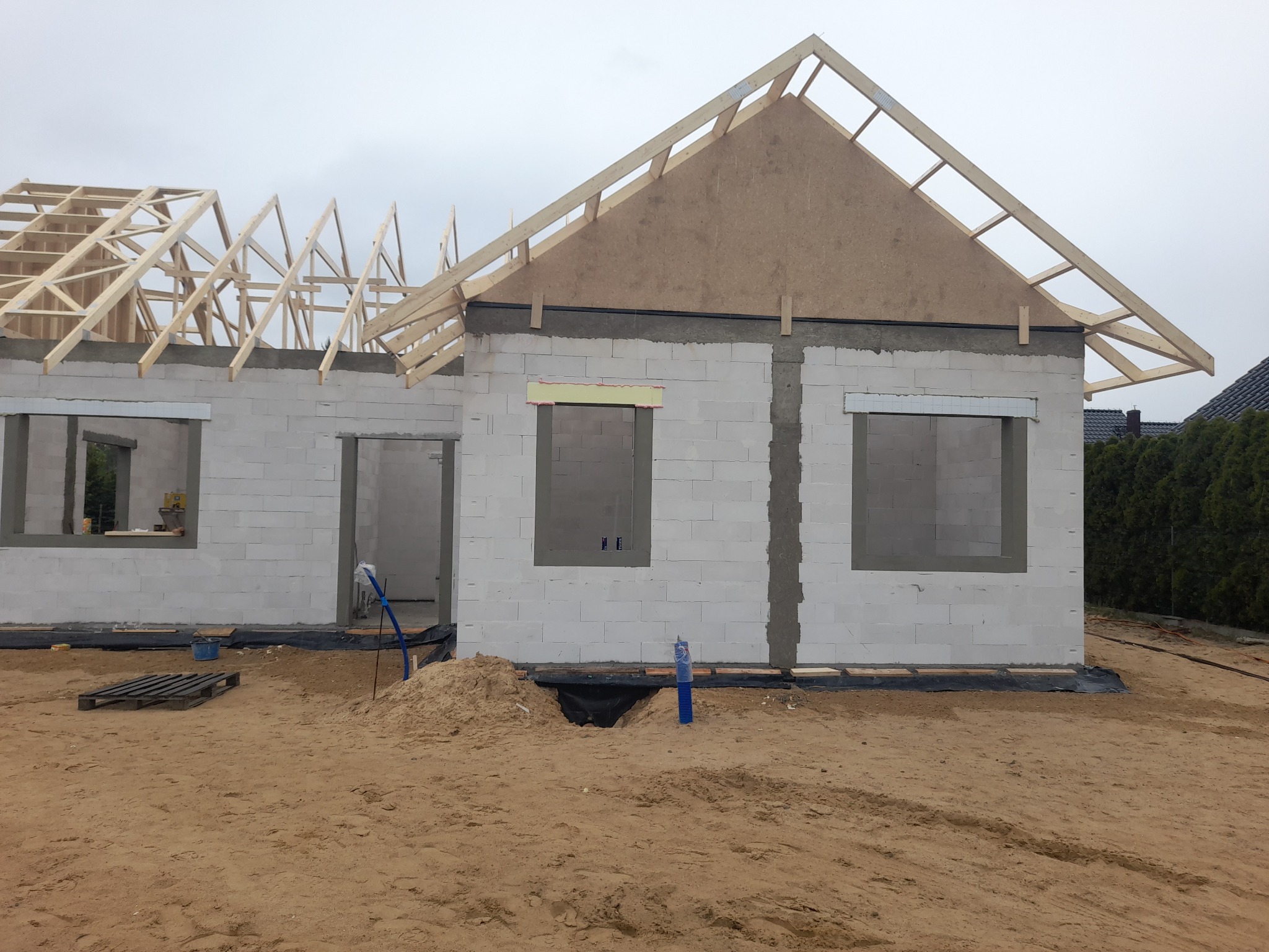 Budowa domu jednorodzinnego w stanie surowym otwartym: białe ściany z betonu komórkowego, drewniana konstrukcja dachu, piasek na działce. Widok z zewnątrz, dzień.