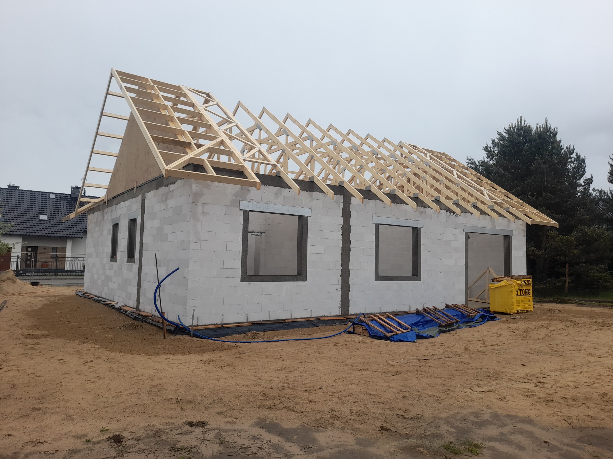 Budowa domu jednorodzinnego w trakcie realizacji: ściany z betonu komórkowego, drewniana konstrukcja dachu, widoczne okna. Na pierwszym planie teren budowy.