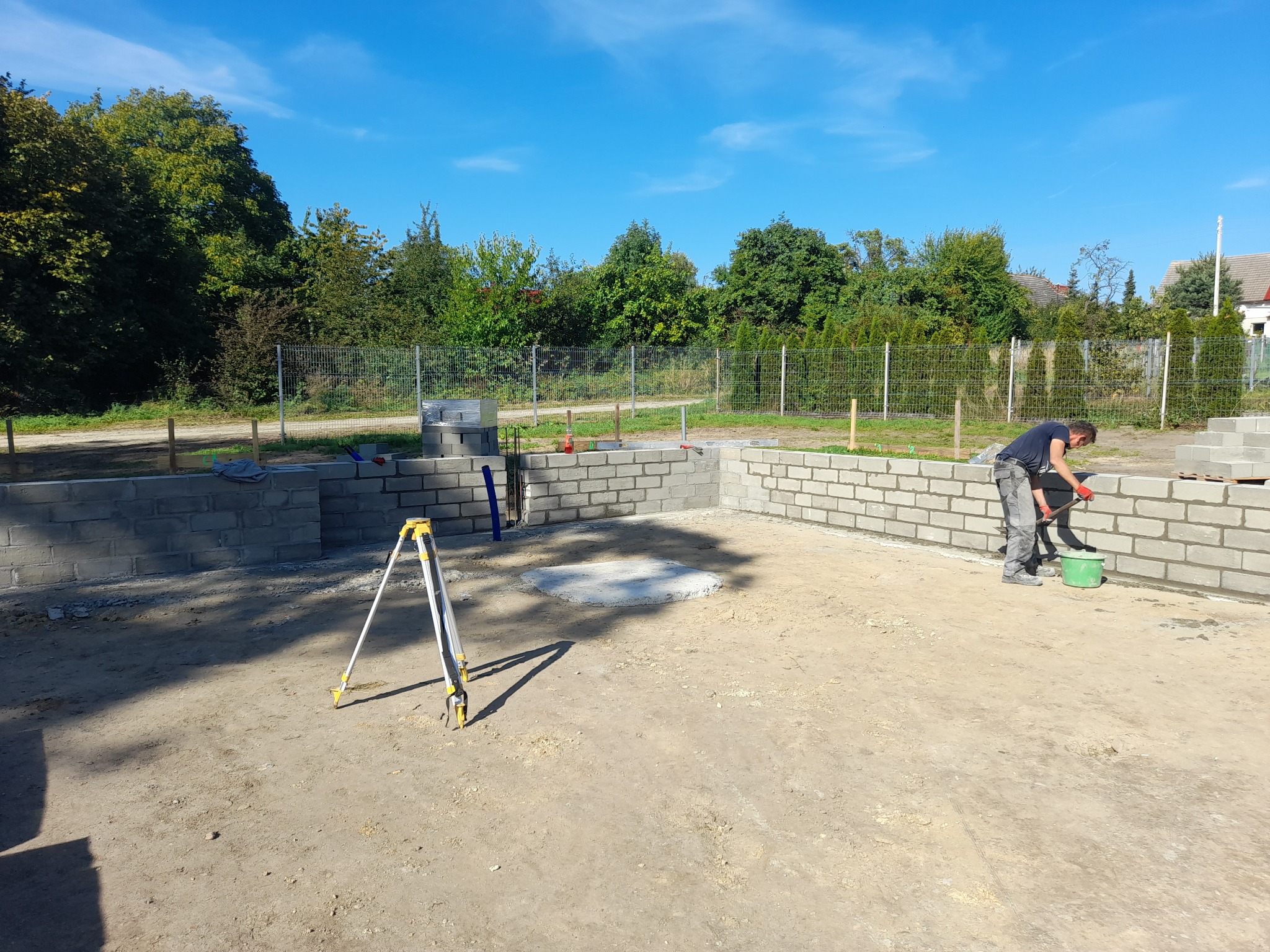 Fundamenty domu w trakcie budowy: ściany z bloczków, pracownik zacierający spoiny, niwelator. Widok na plac budowy w słoneczny dzień.