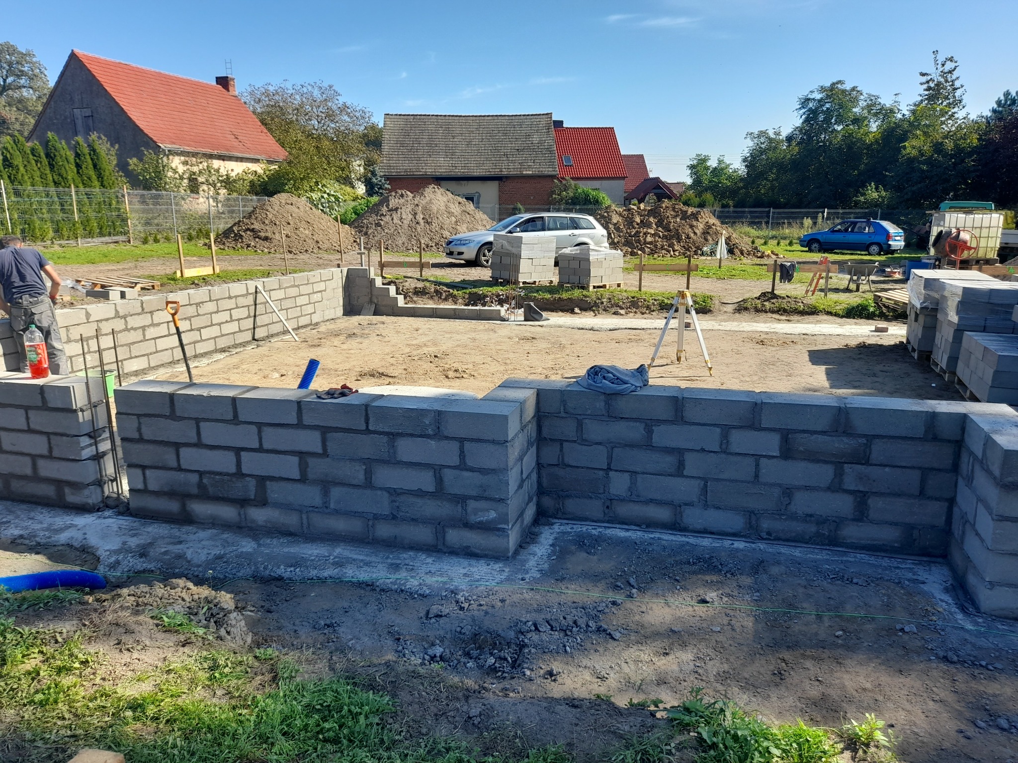 Wznoszenie ścian fundamentowych z bloczków betonowych na budowie domu jednorodzinnego. Widoczny murarz przy pracy, narzędzia i materiały budowlane. Tło: domy i samochody.