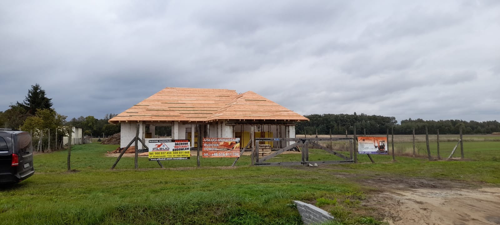 Budowa domu jednorodzinnego w trakcie realizacji. Widoczne ściany z białego bloczka i drewniana konstrukcja dachu. Na ogrodzeniu reklamy firmy budowlanej.