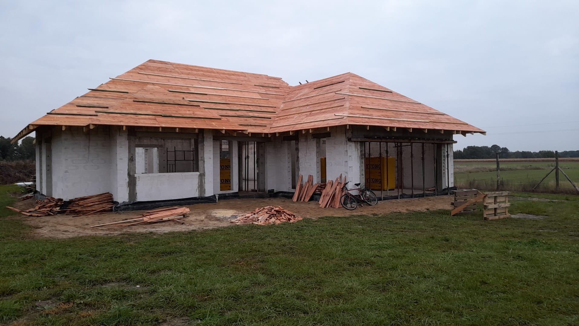 Budowa domu jednorodzinnego z białych bloczków, z częściowo wykonanym drewnianym dachem. Na pierwszym planie leżą deski, a obok stoi rower. Widok na zielone pole.