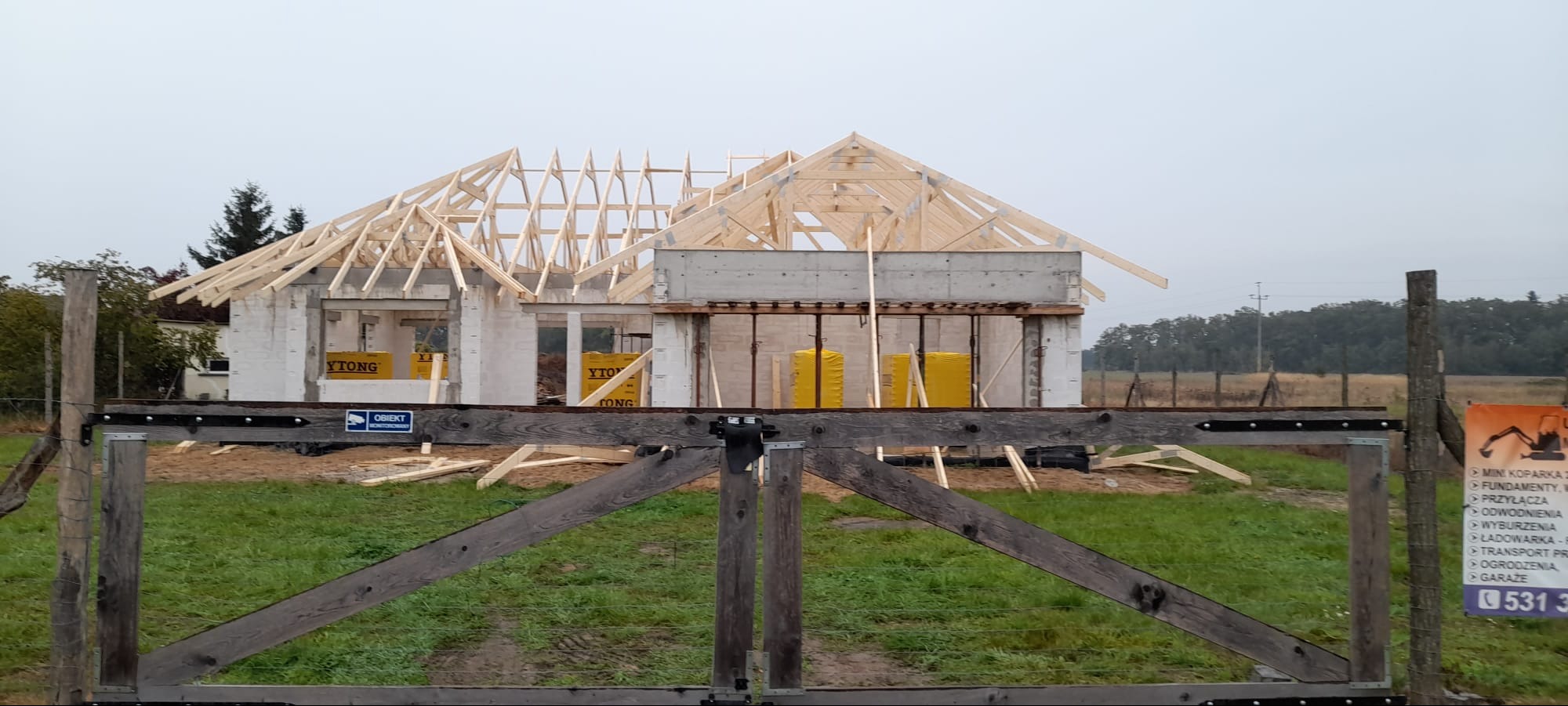 Budowa domu z białego betonu komórkowego Ytong, widoczna konstrukcja dachu z drewna. Ujęcie zza drewnianej bramy wjazdowej na posesję. Tablica reklamowa z boku.