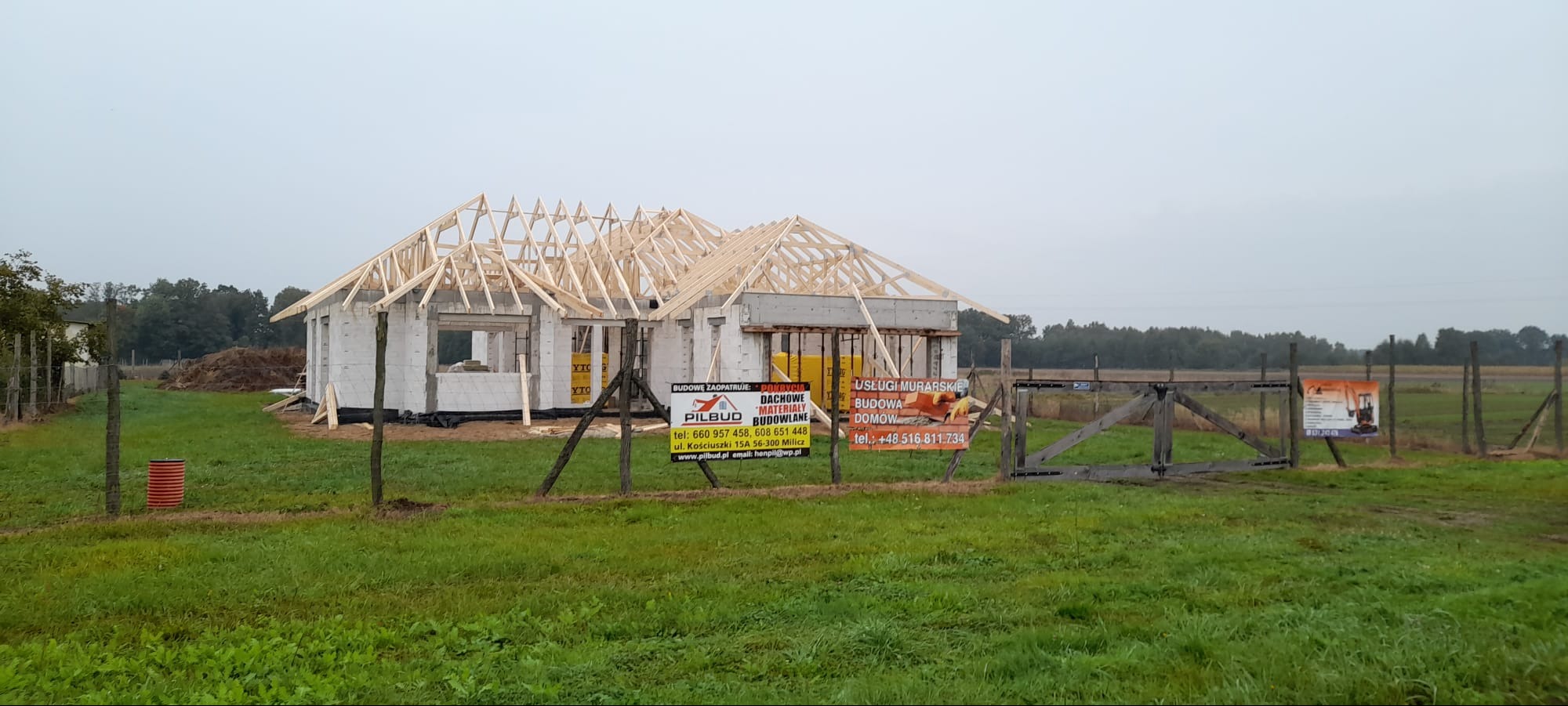 Budowa domu jednorodzinnego w trakcie realizacji: wzniesione ściany z bloczków i konstrukcja dachu. Na pierwszym planie tablice informacyjne firm budowlanych.