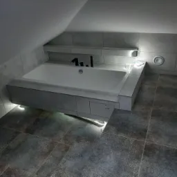 Nowoczesna łazienka na poddaszu z wanną obudowaną płytkami w odcieniach szarości, podświetloną od dołu, ciemna podłoga z wzorzystych płytek, widoczny fragment wentylacji.