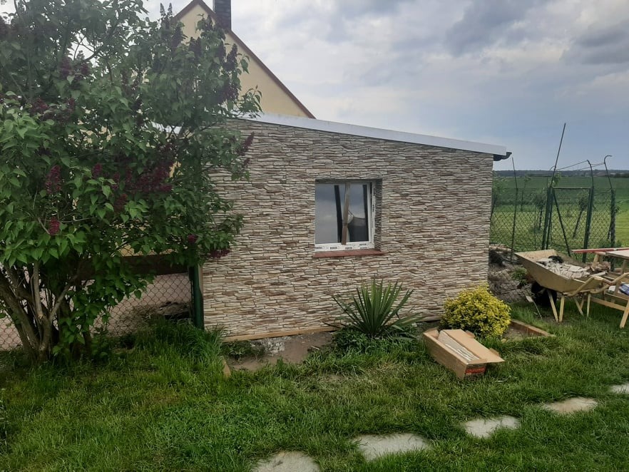 Niewielki budynek gospodarczy z elewacją imitującą kamień, z białym oknem, otoczony zielenią i drzewem kwitnącym na fioletowo, w tle widoczne pole i pochmurne niebo.