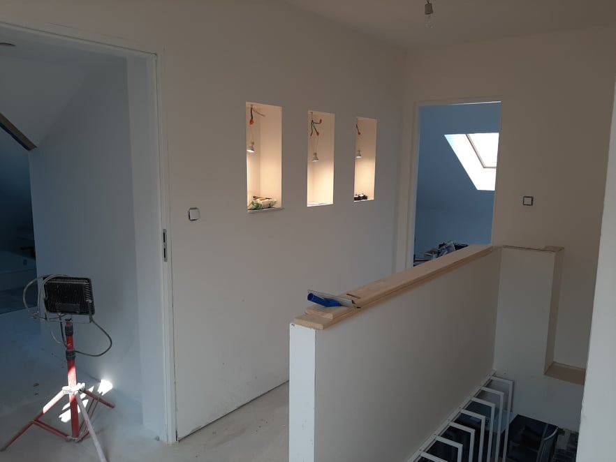 Wnętrze w trakcie remontu: białe ściany z trzema wnękami oświetlonymi punktowo, widoczna lampa budowlana na statywie i schody z metalową balustradą, surowe wykończenie.