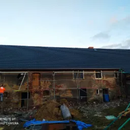 Wymiana dachu na starym budynku z cegły, widoczne rusztowania, pracownik w pomarańczowej kurtce na drabinie, sterta słomy i gruzu na podwórku.