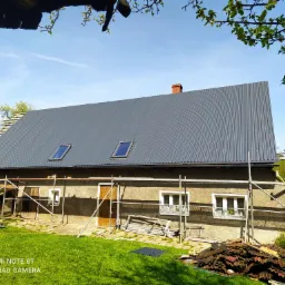 Dom jednorodzinny z nowym, szarym dachem z blachy trapezowej i trzema oknami dachowymi, widoczne rusztowanie i dekarz pracujący na dachu, w tle zielony trawnik i drzewa.