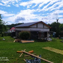 Budynek w trakcie krycia dachu, widoczne rusztowania, materiały budowlane na trawniku, błękitne niebo z chmurami w tle.