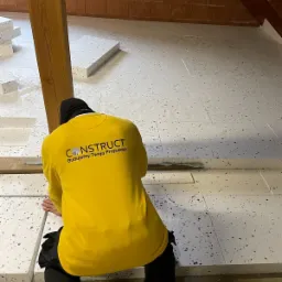 Pracownik w żółtej koszulce z logo firmy układa płyty izolacyjne na poddaszu, widoczna konstrukcja dachu i ceglana ściana.