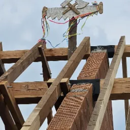 Wieniec ciesielski z dekoracjami na szczycie konstrukcji drewnianej dachu, z widocznymi kominami w trakcie budowy, na tle pochmurnego nieba.