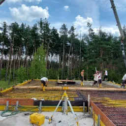 Wylewanie betonu na przygotowany szalunek i zbrojenie fundamentu domu jednorodzinnego, widoczna pompa do betonu Schwung S36X i las w tle.