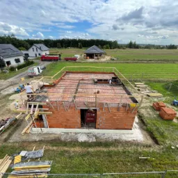 Budowa domu jednorodzinnego z czerwonej cegły, widoczne ściany i przygotowane zbrojenie stropu, w tle zielony krajobraz i zabudowa jednorodzinna.