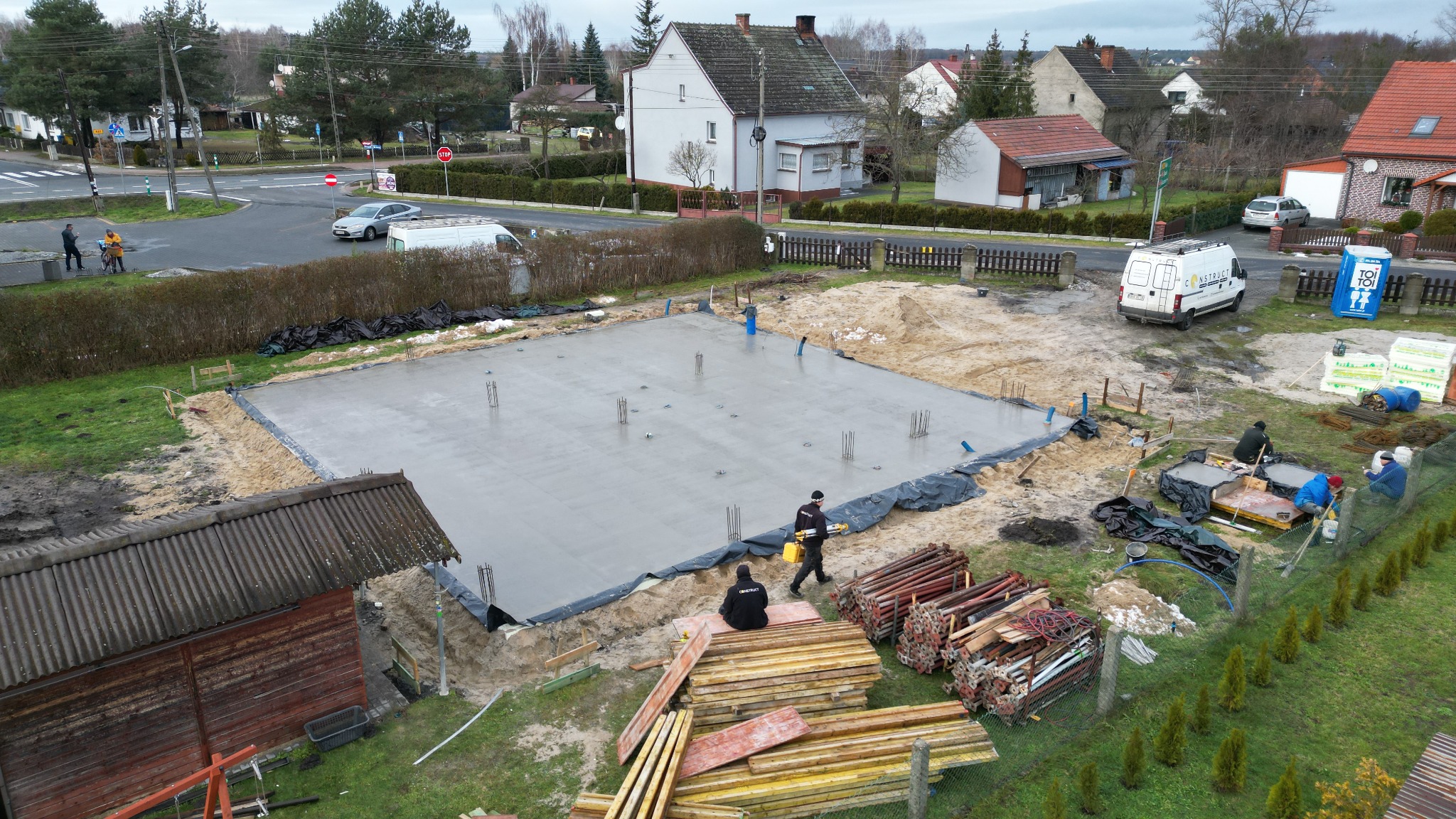 Świeżo wylany fundament domu jednorodzinnego, widoczne wystające pręty zbrojeniowe i ekipa budowlana przy pracy, w tle zabudowa mieszkalna.