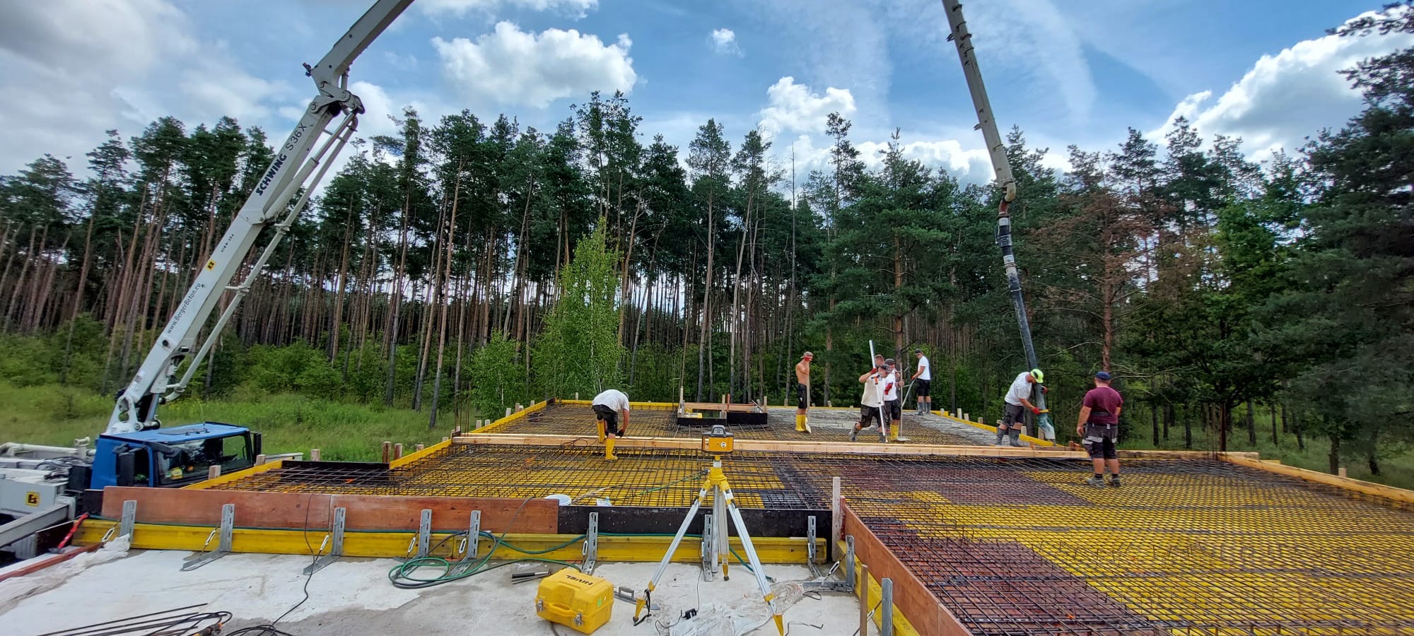 Wylewanie betonu na przygotowany szalunek i zbrojenie fundamentu domu jednorodzinnego, widoczna pompa do betonu Schwung S36X i las w tle.