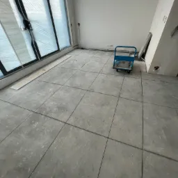 Wnętrze w trakcie remontu z szarymi płytkami podłogowymi, zabezpieczonymi folią oknami i niebieskim wózkiem transportowym.