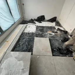 Montaż płytek podłogowych w pomieszczeniu, widoczne czarne folie ochronne, częściowo ułożone płytki w odcieniach szarości i fragment nogi pracownika w spodniach roboczych.