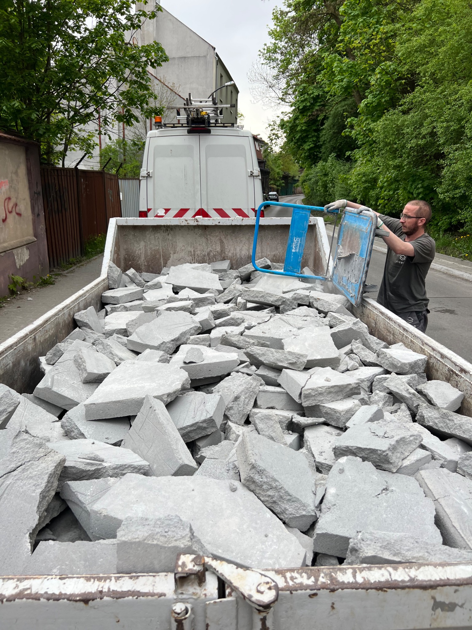 Pracownik w rękawicach wrzuca gruz betonowy do kontenera z widocznym samochodem dostawczym w tle.