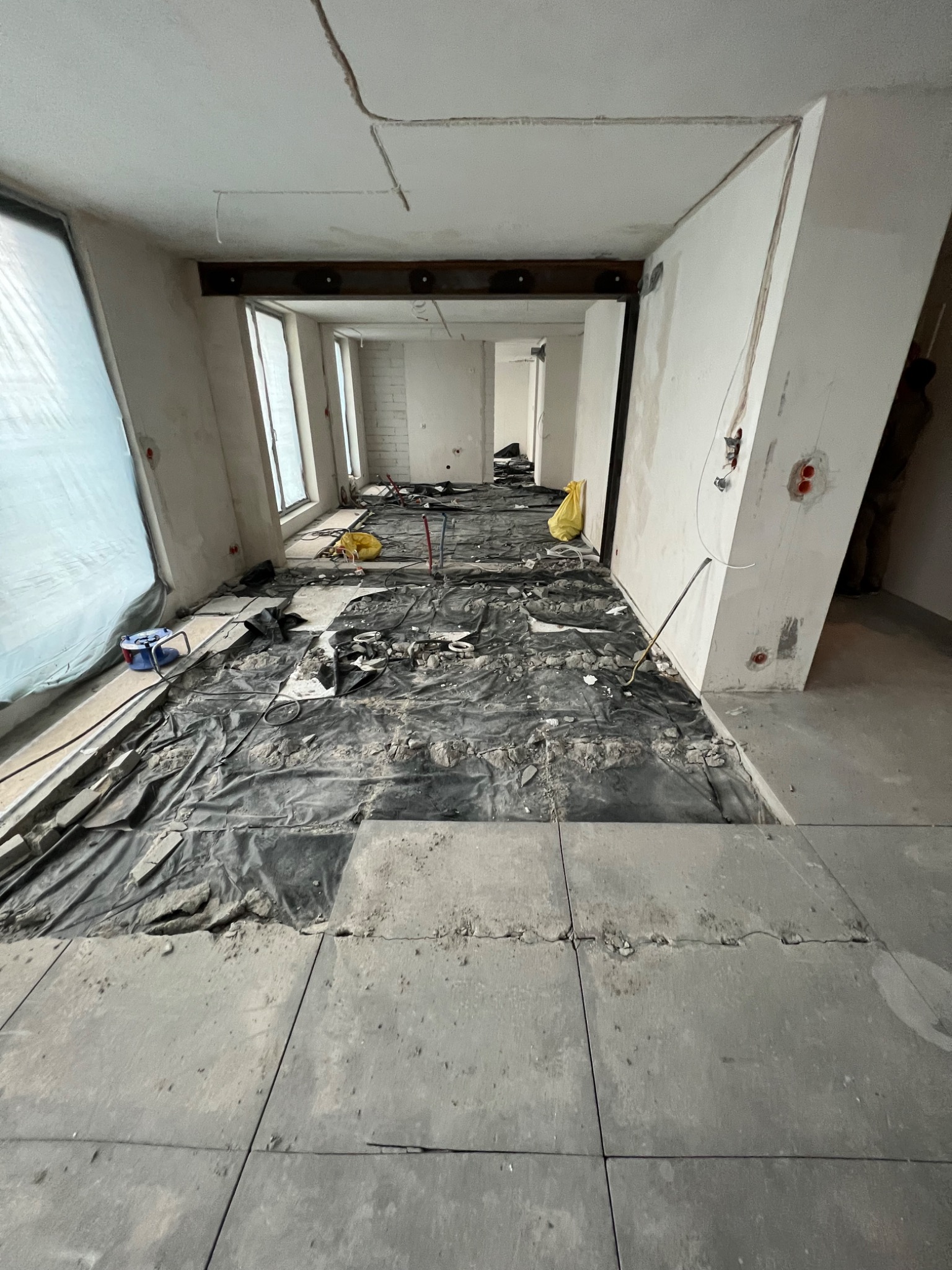 Wnętrze w trakcie remontu, podłoga zabezpieczona czarną folią, widoczne resztki gruzu i narzędzia, surowy stan ścian i stropu z widoczną stalową belką.
