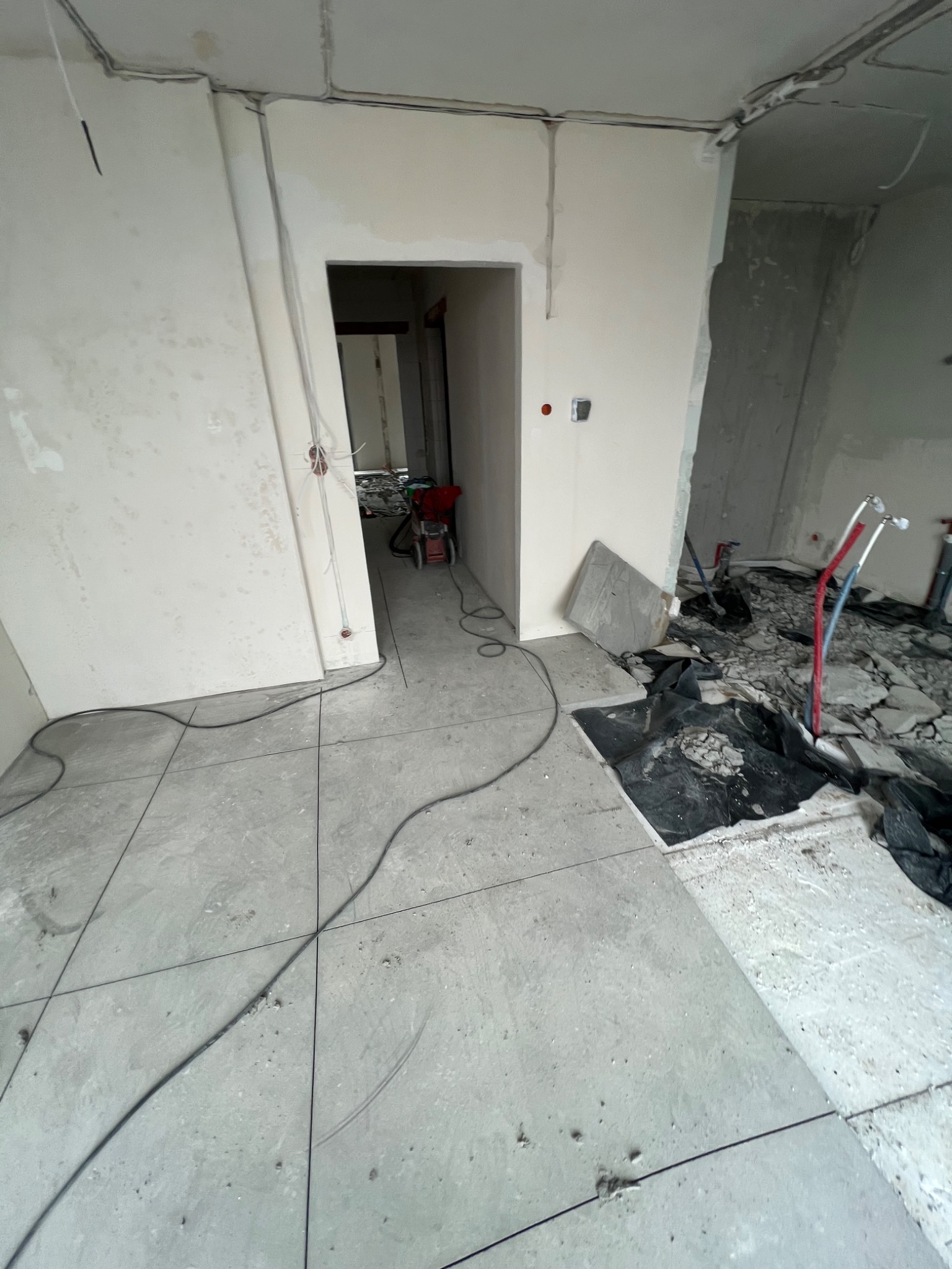 Wnętrze w trakcie remontu: częściowo położona szara podłoga z płytek, odsłonięte przewody elektryczne na ścianach, gruz i folia ochronna w tle, widoczne rury instalacji wodnej, otwór drzwiowy...