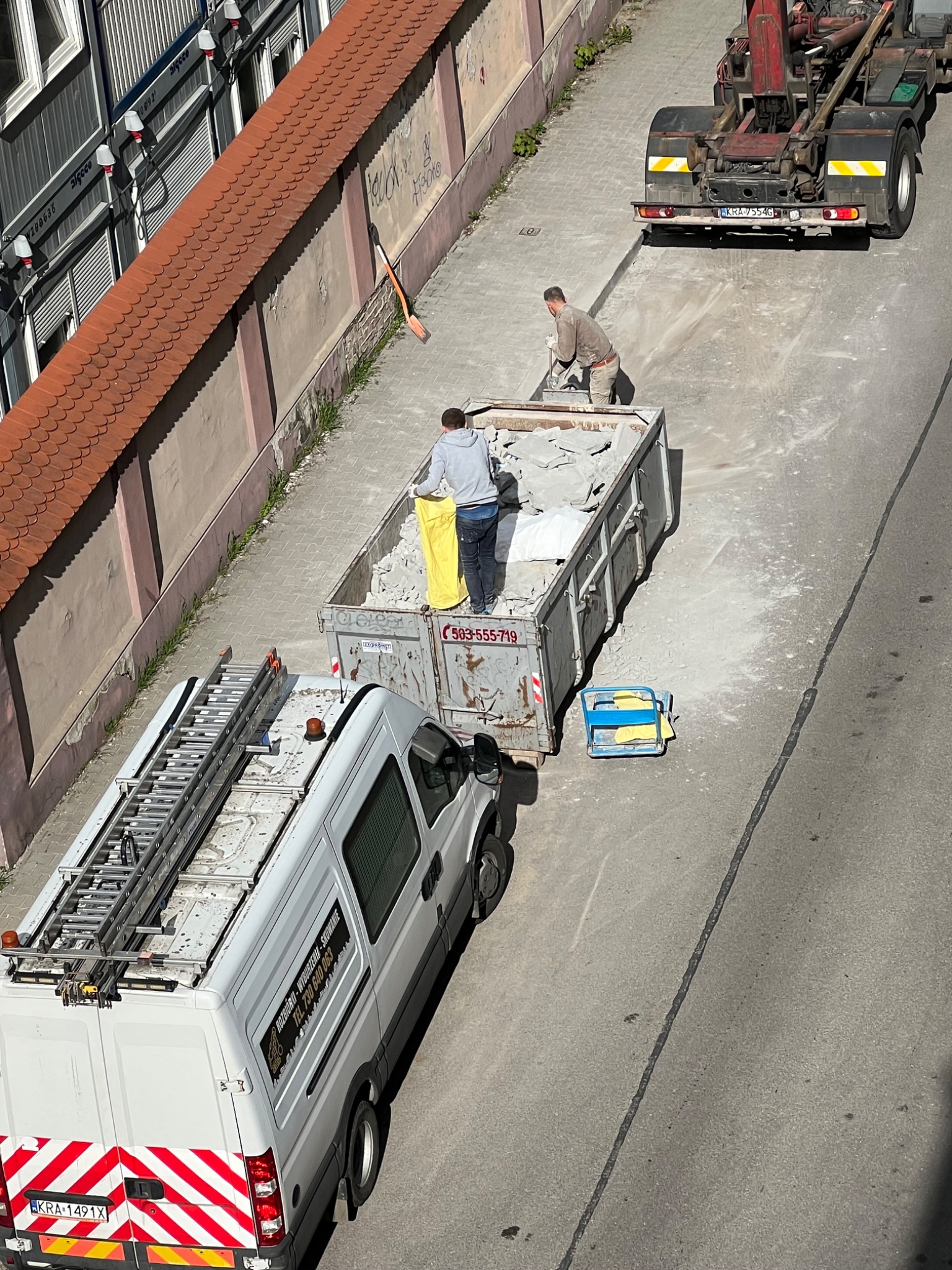 Widok z góry na kontener wypełniony gruzem, dwóch robotników wrzuca do niego kolejne fragmenty, obok stoi biały samochód dostawczy z drabiną na dachu i napisem 'Rozbiórka-Wyburzenia-Skuwanie',...
