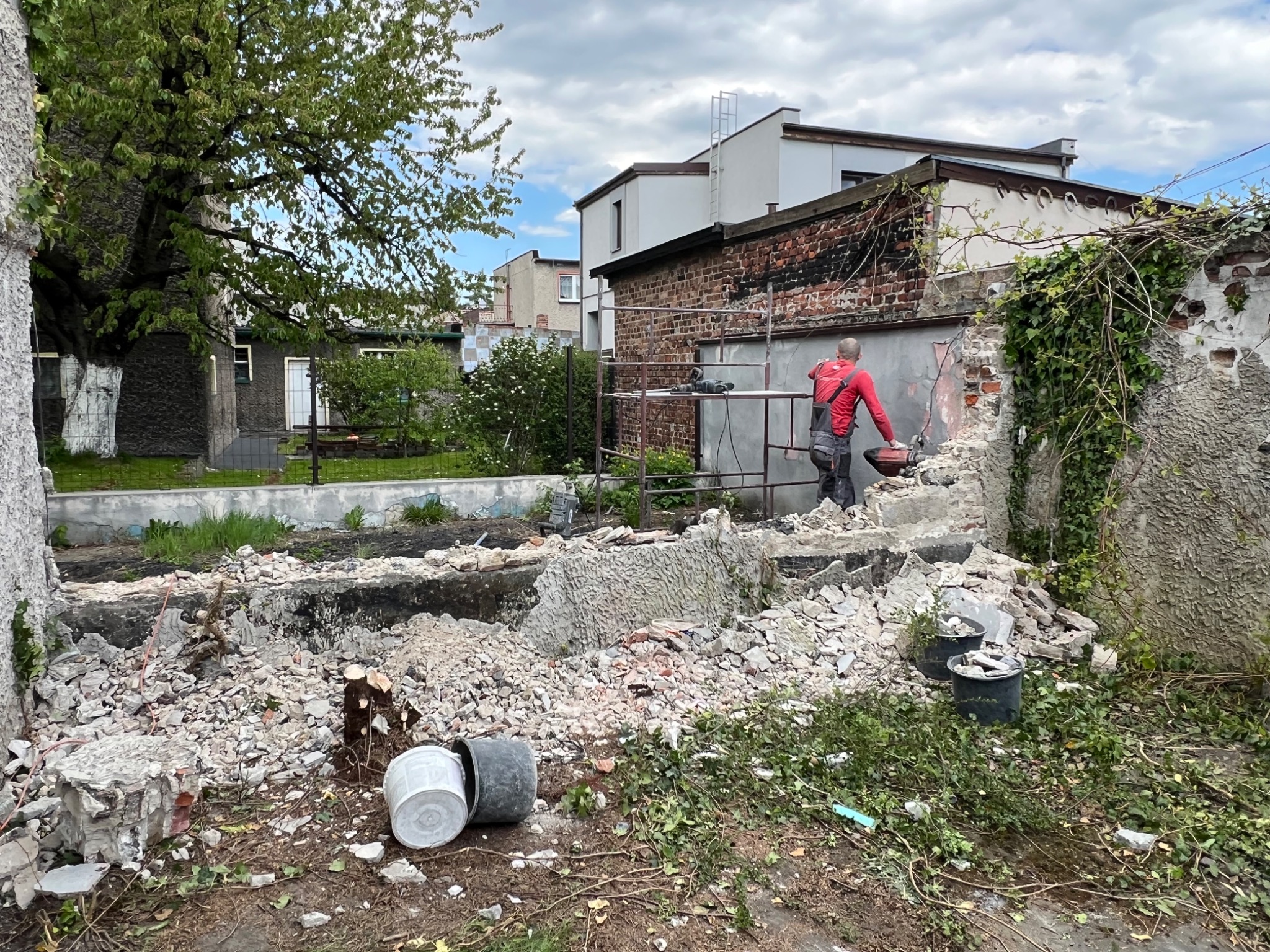 Mężczyzna w czerwonej koszuli na rusztowaniu, z użyciem narzędzia do rozbiórki fragmentu muru, w tle budynki mieszkalne i gruzowisko.