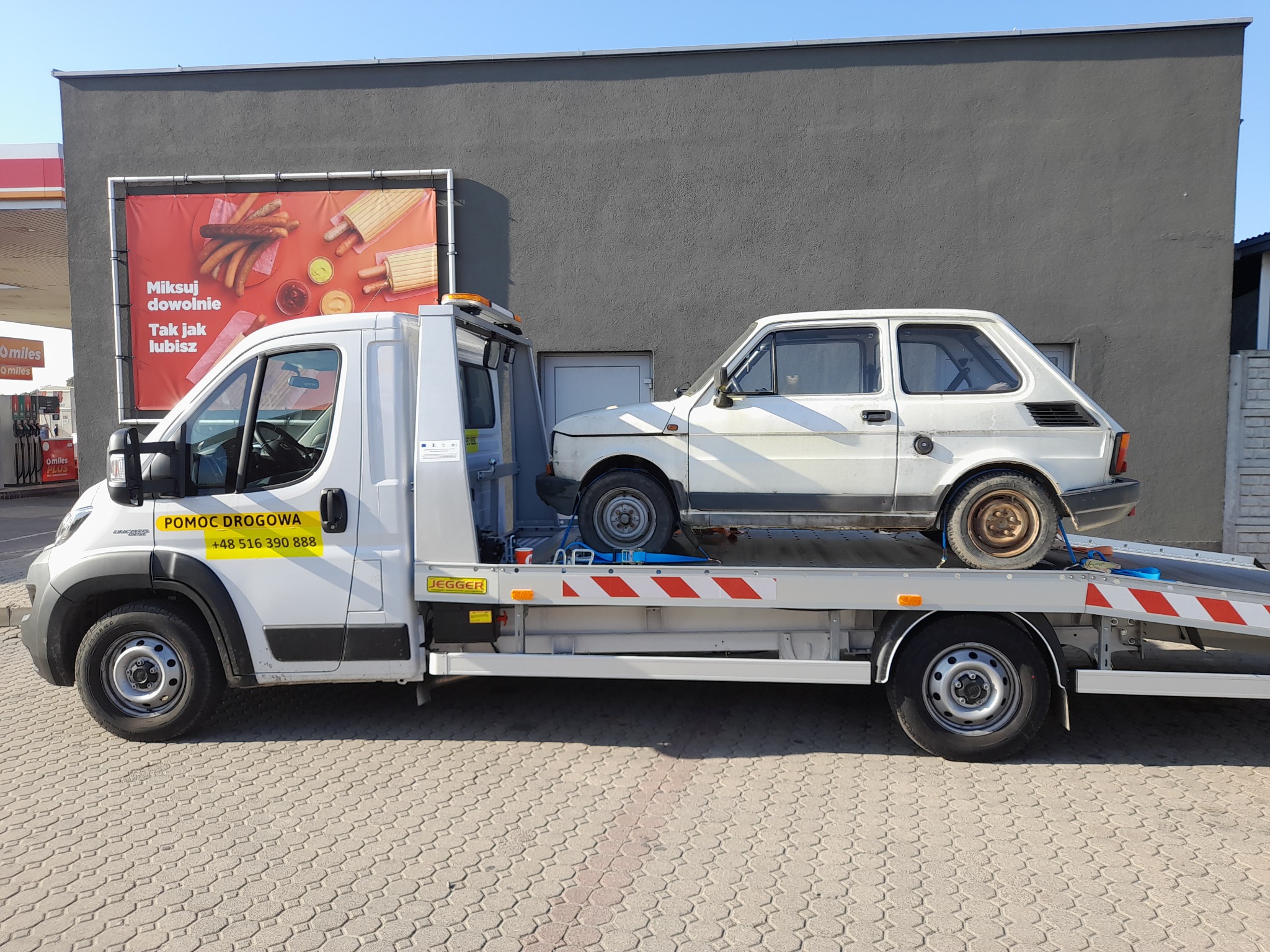 Laweta w Jarosławiu przewozi białego Fiata 126p na tle stacji benzynowej z reklamą jedzenia.