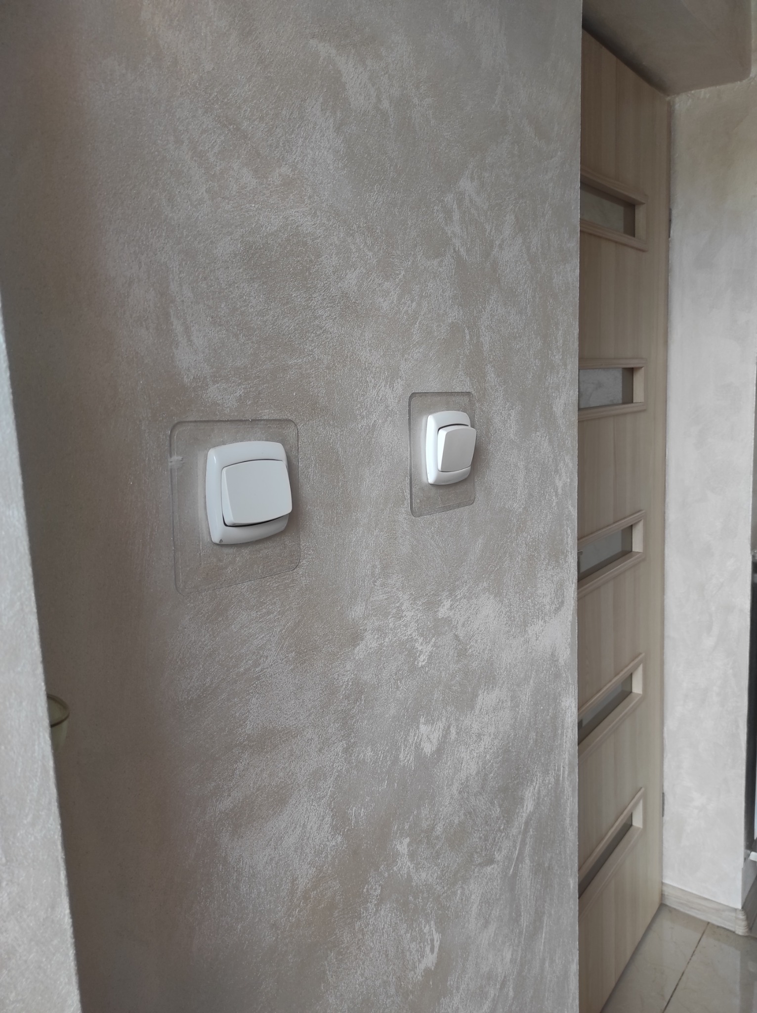 Ściana z dekoracyjną, beżową fakturą, zabezpieczone włączniki światła i fragment drewnianych drzwi z przeszkleniami.