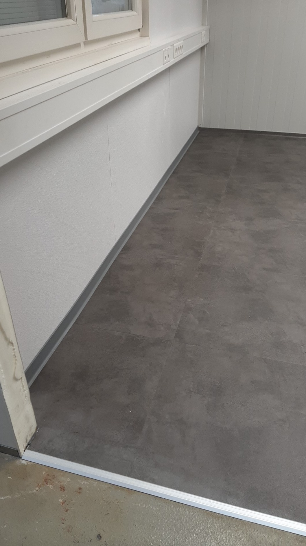 Szare panele podłogowe imitujące kamień, ułożone w pomieszczeniu biurowym z białymi ścianami i listwami przypodłogowymi, widoczne gniazdka elektryczne pod oknem.