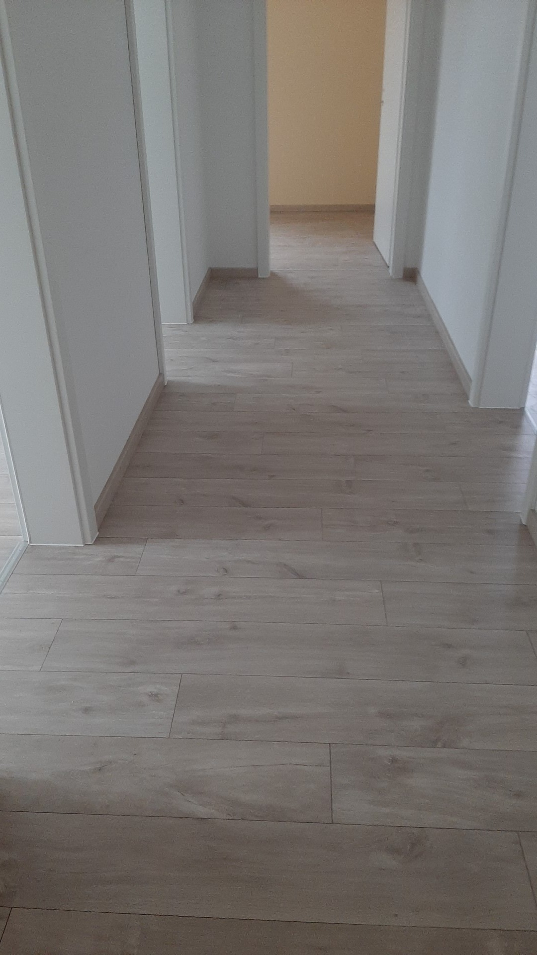 Jasne panele podłogowe ułożone wzdłuż długiego korytarza z białymi ścianami i otwartymi drzwiami do sąsiednich pomieszczeń.