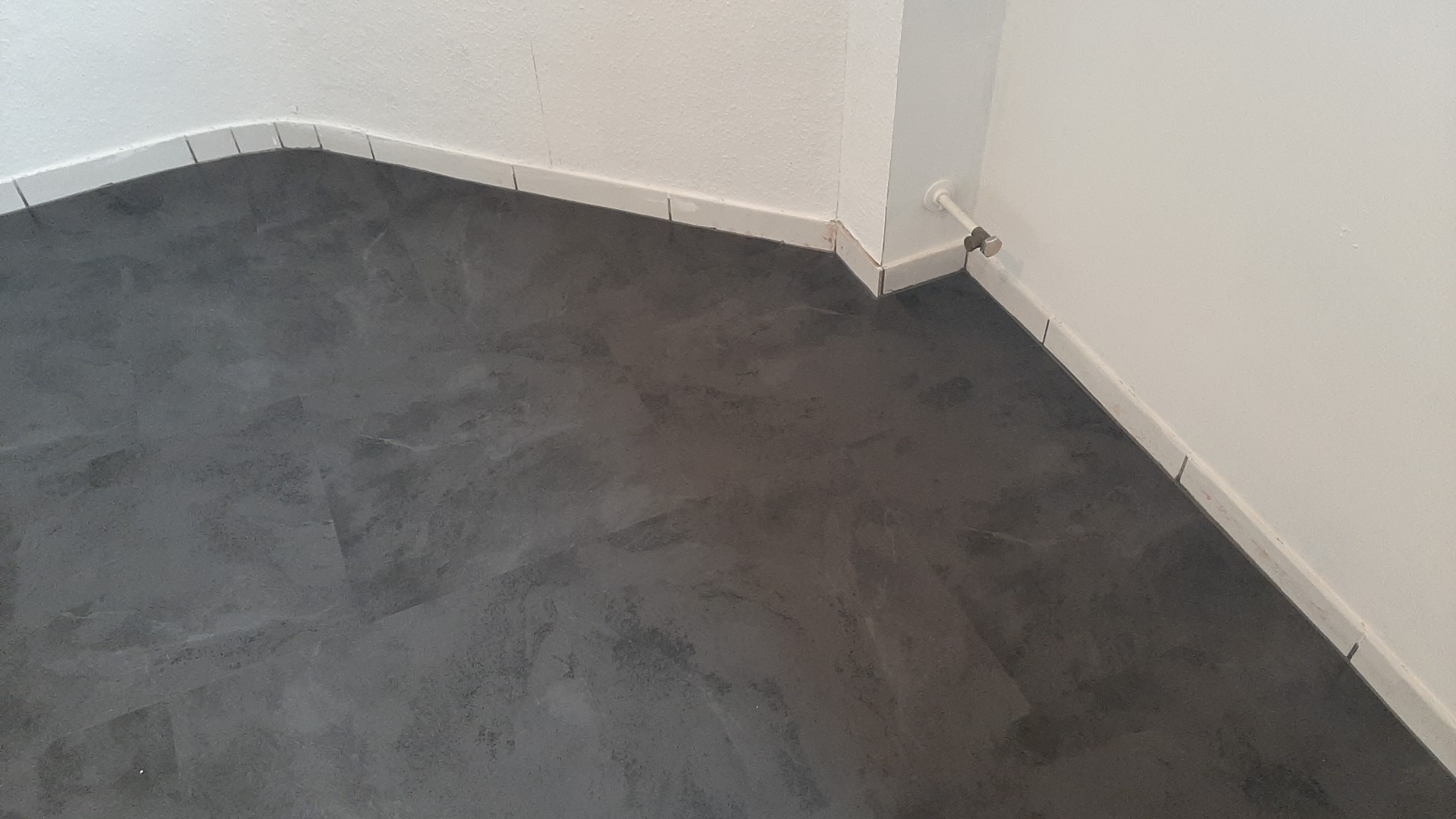 Ułożona, ciemnoszara podłoga z paneli w narożniku pomieszczenia, z białą listwą przypodłogową i rurą grzewczą na ścianie.