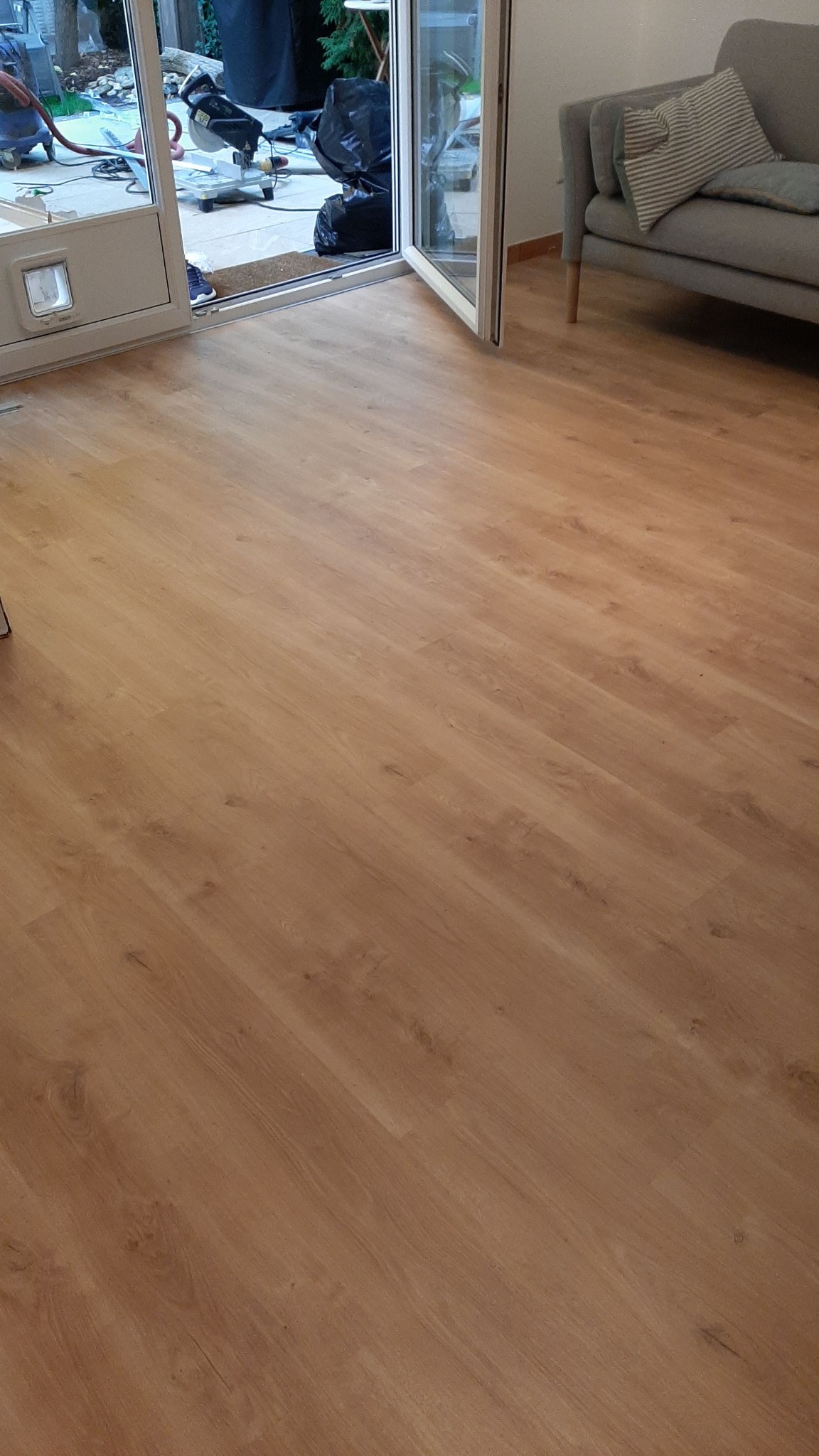 Ułożona podłoga z paneli w odcieniach brązu, widoczny fragment sofy i otwarte drzwi balkonowe z narzędziami na zewnątrz.