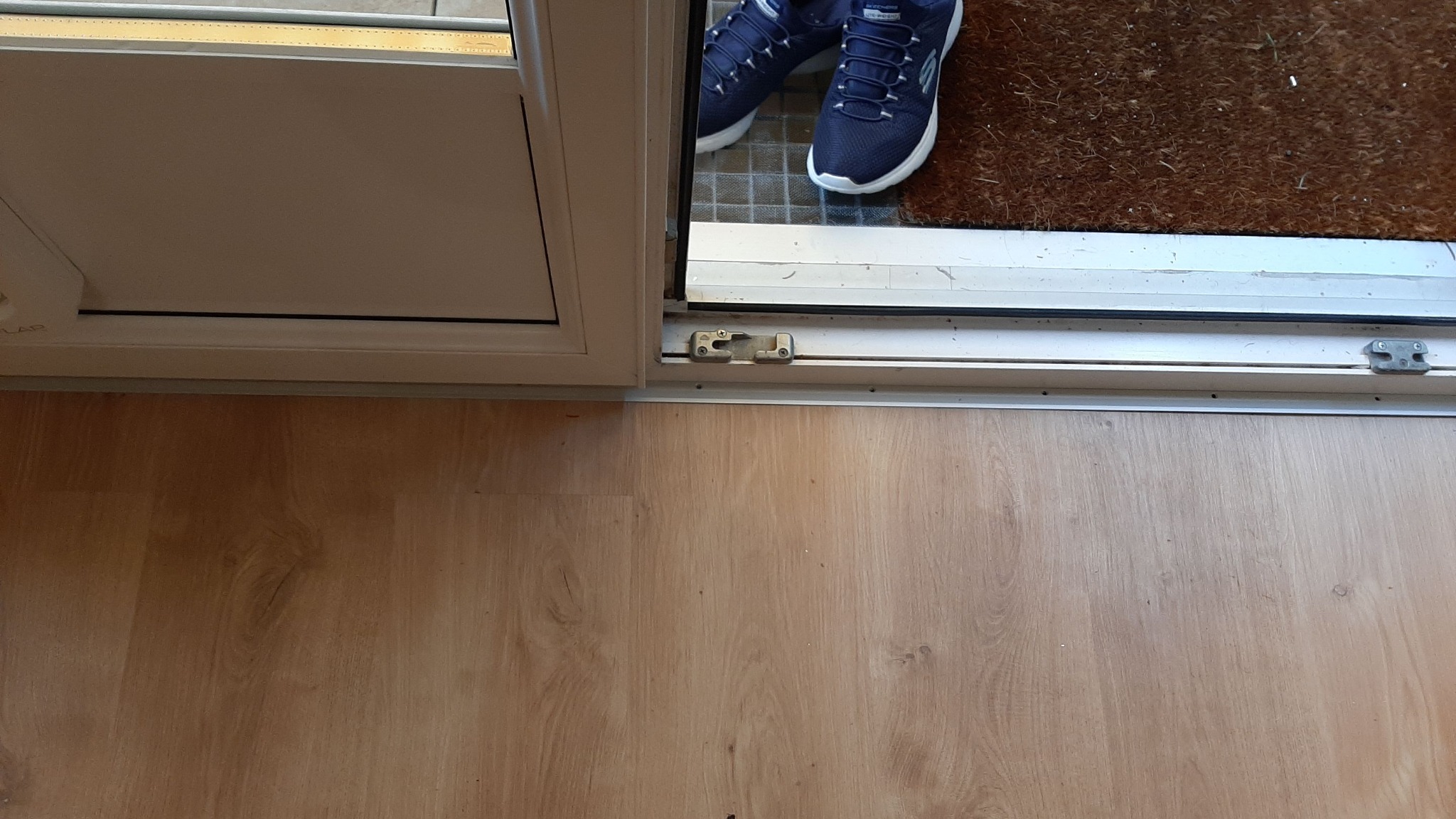 Fragment podłogi z paneli, widoczny próg drzwi balkonowych, fragment wycieraczki i pary granatowych butów sportowych na niej.