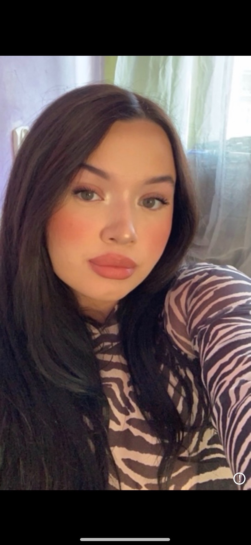 Portret młodej kobiety z długimi, ciemnymi włosami i makijażem, ubrana w bluzkę w zebrę, pozująca na tle okna z jasnymi zasłonami.